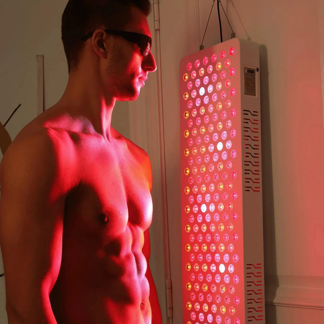 Man wearing protective goggles beside a wall-mounted red light therapy panel glowing red during a session